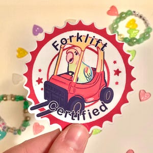 May include: A sticker featuring a cartoon parrot driving a red forklift. The sticker is circular with a red and white scalloped edge. Text on the sticker reads "Forklift Certified".