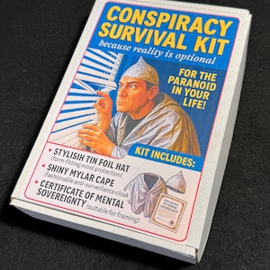 May include: A white box labeled "CONSPIRACY SURVIVAL KIT" with a blue and yellow graphic. The kit includes a tin foil hat, a shiny mylar cape, and a certificate of mental sovereignty. The box features an illustration of a man wearing a tin foil hat.