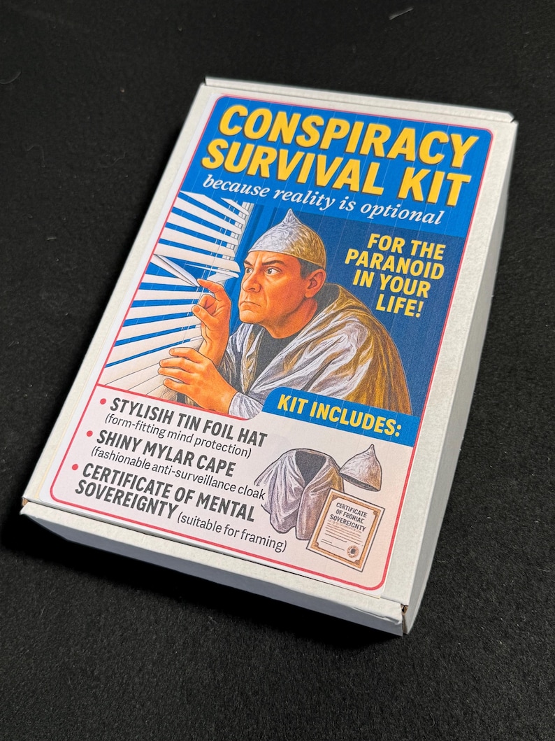 May include: A white box labeled "CONSPIRACY SURVIVAL KIT" with a blue and yellow graphic. The kit includes a tin foil hat, a shiny mylar cape, and a certificate of mental sovereignty. The box features an illustration of a man wearing a tin foil hat.