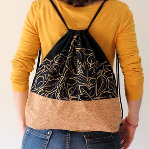 May include: A black drawstring backpack with a gold leaf pattern and a cork bottom. The backpack is being worn by a person.