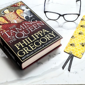 May include: A book titled "The Taming of the Queen" by Philippa Gregory. The book is a paperback with a red and black cover featuring a portrait of a king and queen. A yellow and black bee-themed bookmark is placed on the book, and a pair of black-framed glasses is lying on a white marble surface.