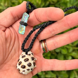 May include: A necklace featuring a white seashell pendant with dark brown spots, suspended from a black braided cord. The cord includes an iridescent abalone shell toggle closure, showcasing vibrant blue, green, and purple hues in a rectangular shape.
