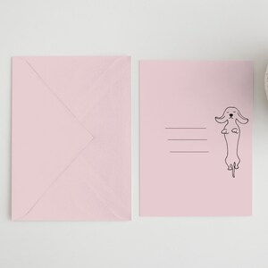 May include: A pink envelope with a blank pink card featuring a black line drawing of a dachshund standing on its hind legs.