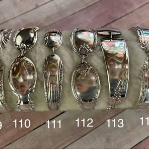 May include: Six silver spoon bracelets, each with a different pattern. The bracelets are shown on a light grey surface, with the numbers 109, 110, 111, 112, 113, and 114 visible below each bracelet.