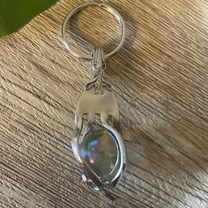 Könnte beinhalten: Ein handgefertigter, silberfarbener Schlüsselanhänger mit einem Design aus einer wiederverwendeten Gabel. Die Zinken der Gabel halten einen schimmernden, irisierenden Stein. Der Schlüsselanhänger hat einen Metallring zum Befestigen von Schlüsseln. Holzuntergrund.