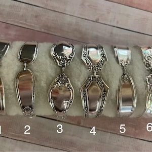 May include: Six silver spoon pendants with different designs. Each pendant is made from a repurposed spoon, with the spoon bowl forming the main body of the pendant. The designs include floral, geometric, and ornate patterns. The pendants are displayed on a white surface.
