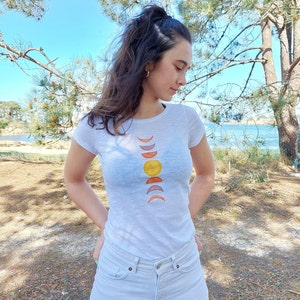 May include: A white t-shirt with a graphic of a sun and moon phases in shades of orange, yellow, and pink.