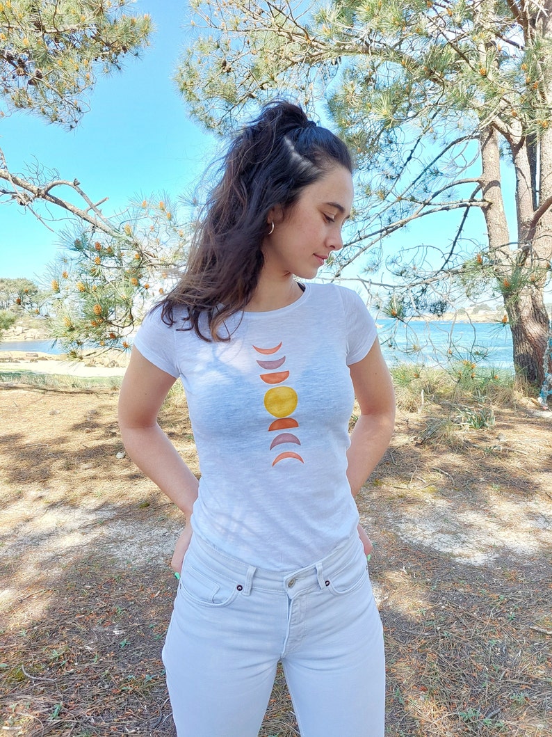 May include: A white t-shirt with a graphic of a sun and moon phases in shades of orange, yellow, and pink.
