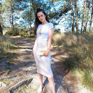 May include: A woman wearing a white t-shirt with a moon and sun graphic and a white skirt. She is holding a white clutch with a moon and sun graphic. She is standing on a dirt path in a forest.