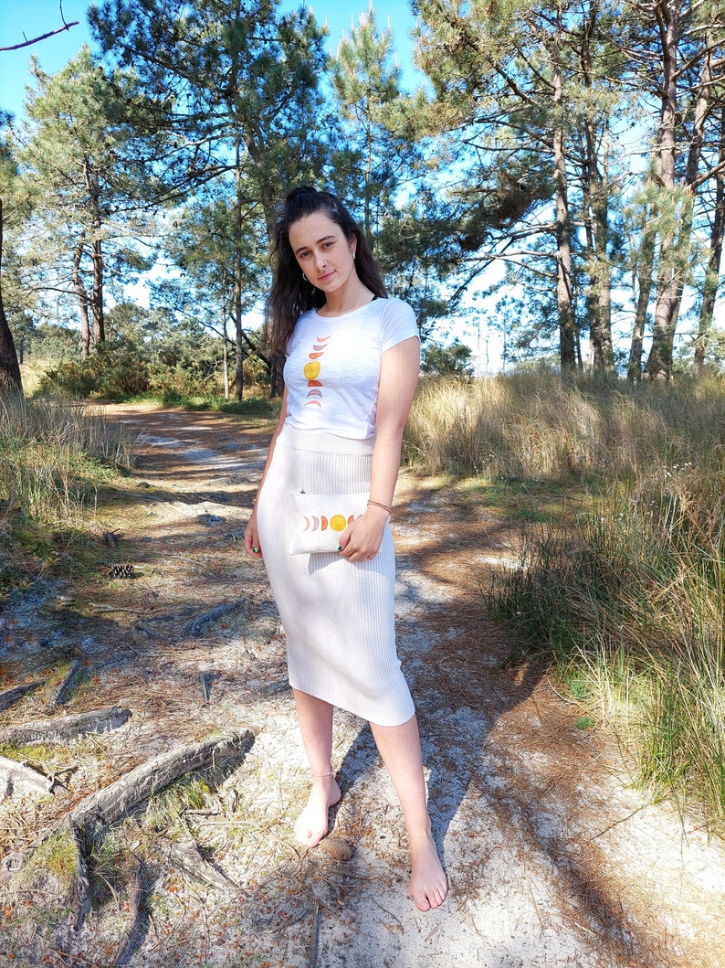 May include: A woman wearing a white t-shirt with a moon and sun graphic and a white skirt. She is holding a white clutch with a moon and sun graphic. She is standing on a dirt path in a forest.