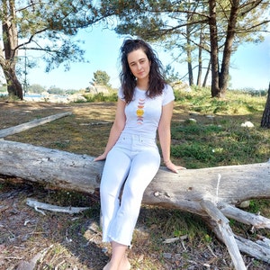 May include: A woman wearing a white t-shirt with a graphic of a sun and moon phases design sits on a fallen tree trunk in a forest setting.