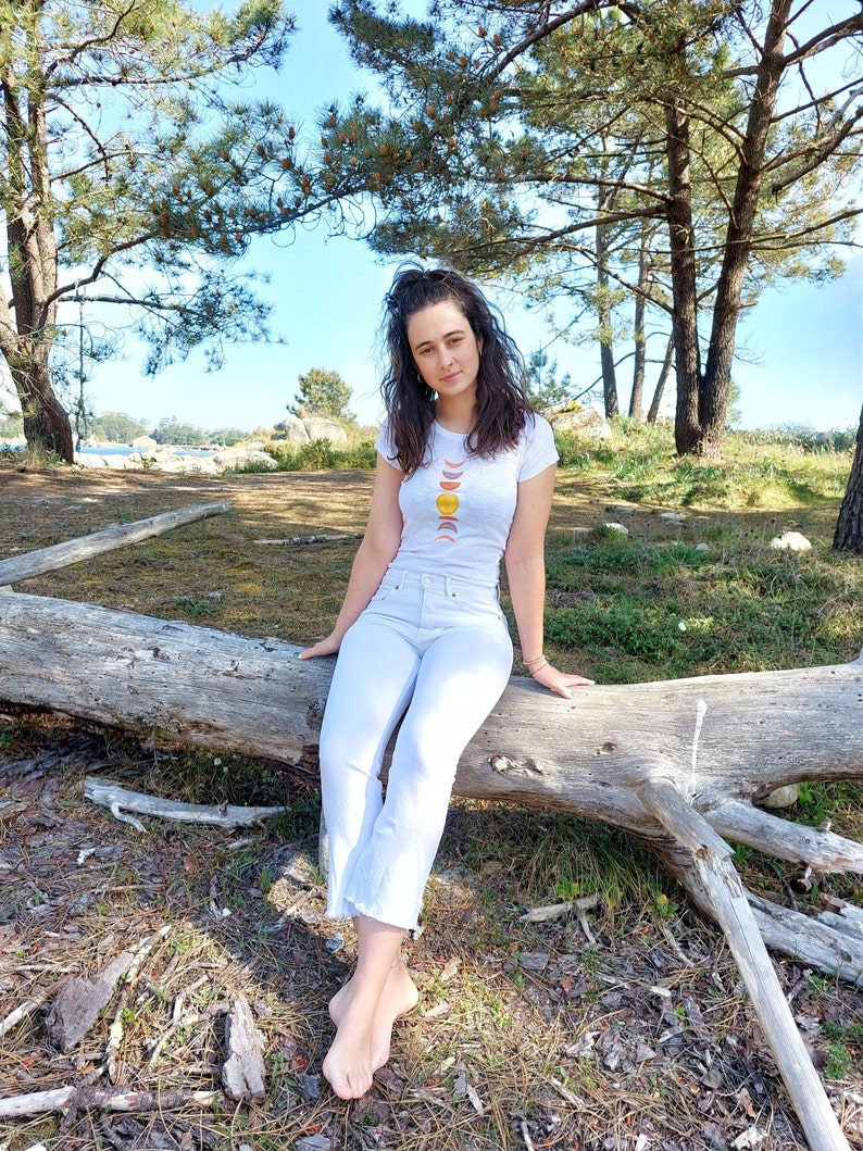 May include: A woman wearing a white t-shirt with a graphic of a sun and moon phases design sits on a fallen tree trunk in a forest setting.