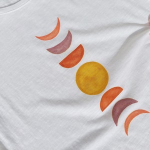 May include: White t-shirt with a design of a yellow sun and orange, red, and purple crescent moons.