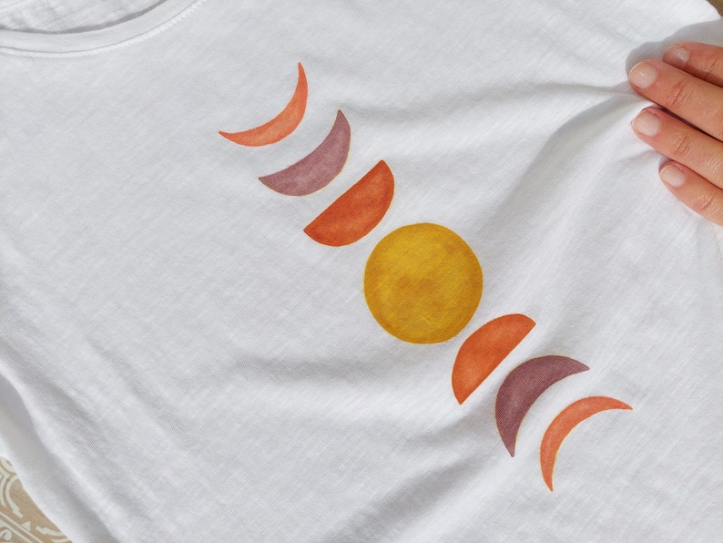 May include: White t-shirt with a design of a yellow sun and orange, red, and purple crescent moons.