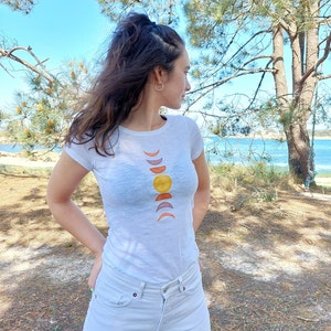 May include: A white t-shirt with a graphic design of a sun and crescent moons in shades of orange, yellow, and brown.