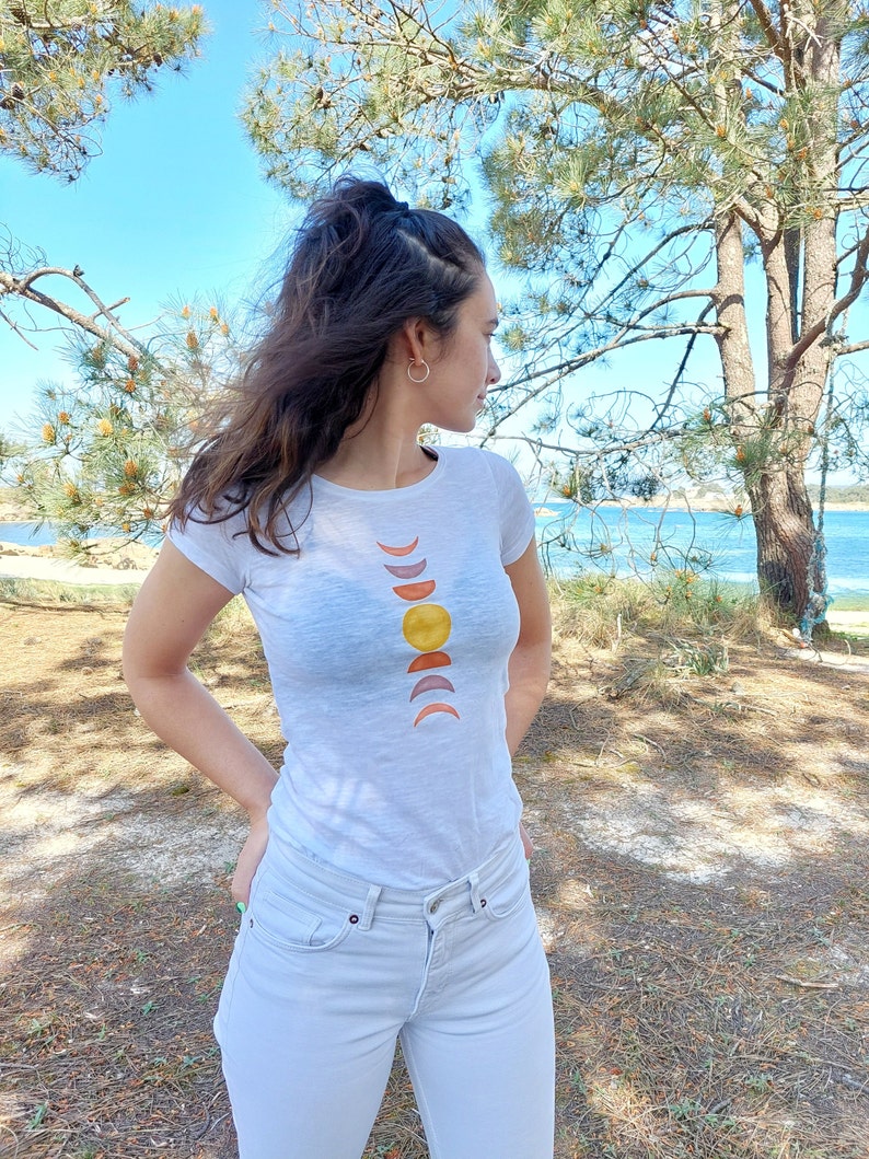 May include: A white t-shirt with a graphic design of a sun and crescent moons in shades of orange, yellow, and brown.
