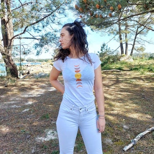 May include: A white t-shirt with a graphic of a sun and moon phases in shades of yellow, orange, and red. The person wearing the shirt is standing in a forest setting.