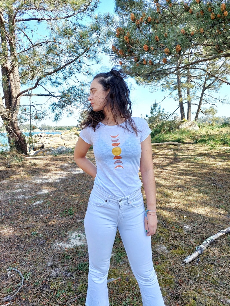 May include: A white t-shirt with a graphic of a sun and moon phases in shades of yellow, orange, and red. The person wearing the shirt is standing in a forest setting.