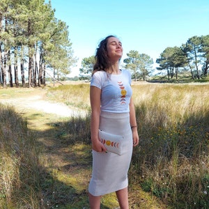 May include: A woman wearing a white t-shirt with a moon and sun phases design and a beige skirt. She is holding a beige clutch with a similar moon and sun phases design. She is standing in a field with tall grass and trees in the background.