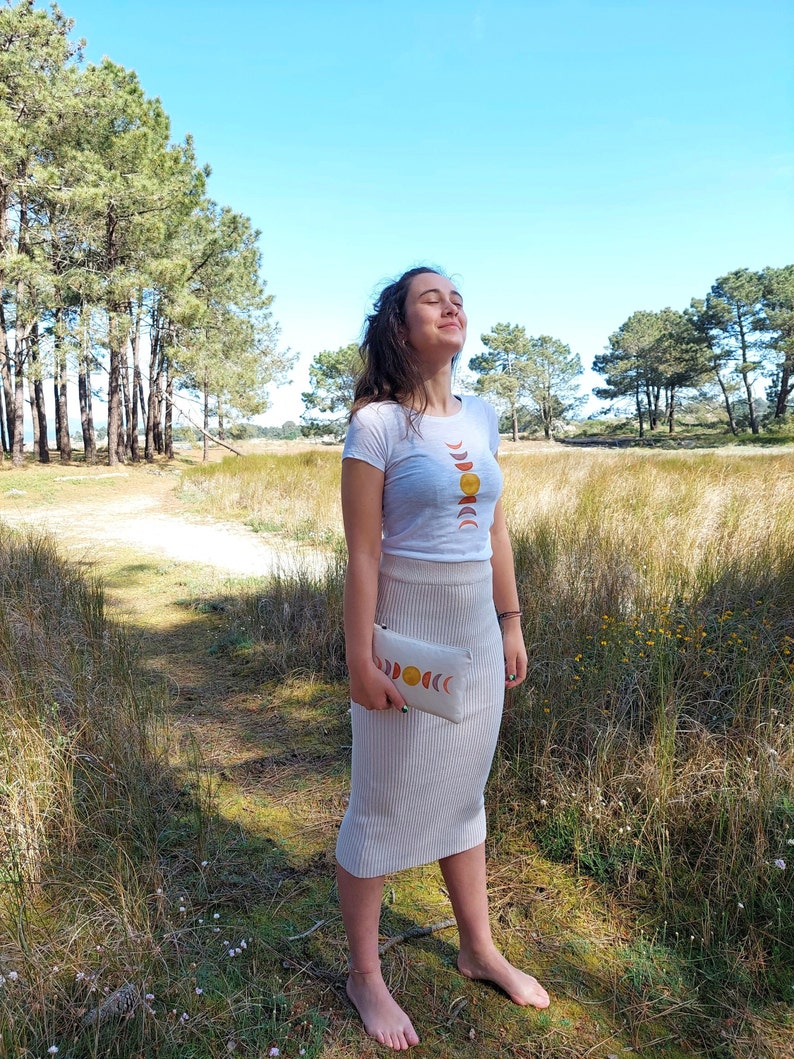 May include: A woman wearing a white t-shirt with a moon and sun phases design and a beige skirt. She is holding a beige clutch with a similar moon and sun phases design. She is standing in a field with tall grass and trees in the background.