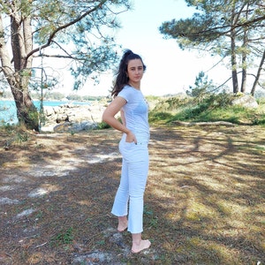 May include: A young person wearing a white t-shirt and white jeans stands in a grassy area with trees in the background.