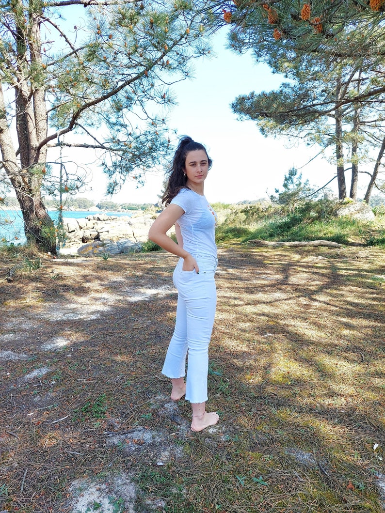 May include: A young person wearing a white t-shirt and white jeans stands in a grassy area with trees in the background.
