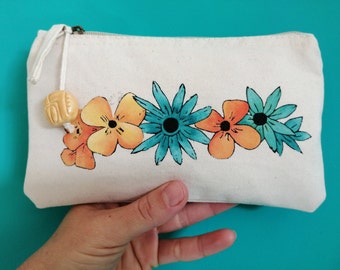 May include: A white canvas zippered pouch with a hand-painted design of orange and yellow flowers and a teal blue flower. The pouch has a brown zipper and a small beige tassel.