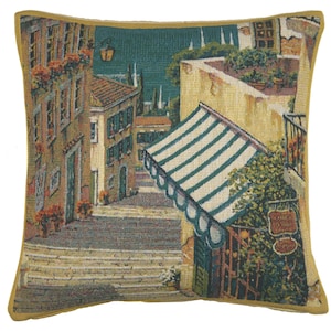 May include: Decorative square pillow with a woven tapestry design depicting a European street scene. The pillow features buildings with green shutters, a striped awning, and a sign that reads "CONCA D'ORO". The pillow has a yellow border.