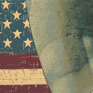 May include: A close-up of a tapestry featuring a faded American flag with red, white and blue stripes and yellow stars. The flag is partially obscured by a textured, beige background.