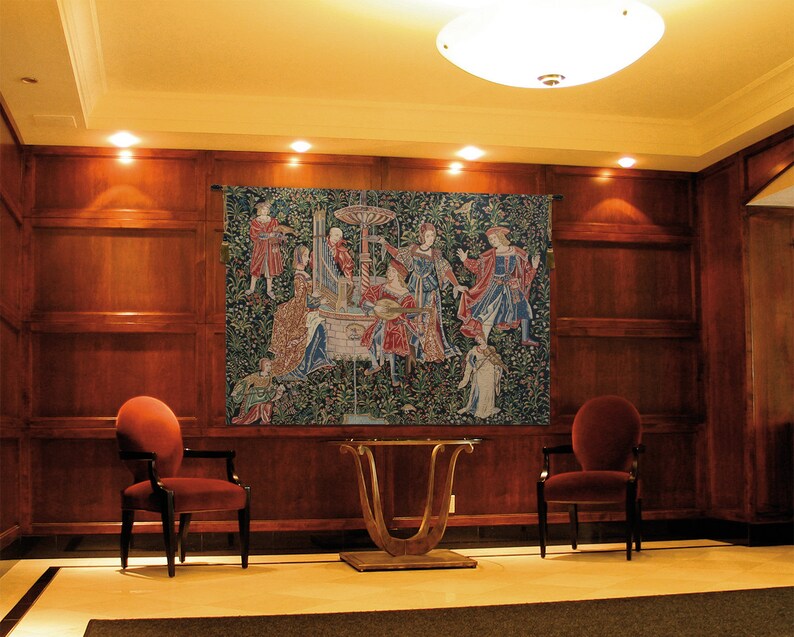 May include: A tapestry with a medieval scene of people dancing and playing music in a garden setting. The tapestry is hanging on a wall in a room with wood paneling and two red chairs. The tapestry has a green background with red, blue, and yellow accents.
