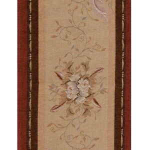 May include: A beige table runner with a brown and red border. The runner has a floral design with pink and white flowers and leaves. The runner has tassels on each end.