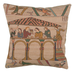 May include: A tapestry pillow featuring a scene from the Bayeux Tapestry, depicting a group of people seated at a table under a canopy. The scene is set against a background of green and brown, with birds and other details woven into the fabric. The pillow is made of a soft, beige fabric and has a zipper closure.