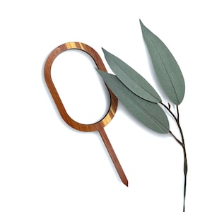 May include: A wooden cake topper with an oval frame and a pointed handle. The topper is made of light brown wood with a natural grain pattern. Next to it is a sprig of faux eucalyptus leaves in shades of green and gray.