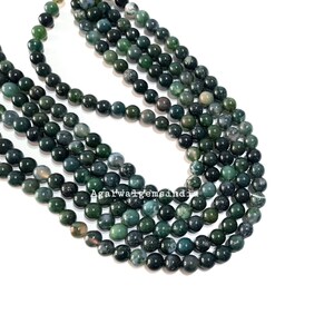 May include: A strand of green moss agate beads, each bead is round and smooth. The beads are arranged in a circular pattern, with the ends of the strand overlapping.