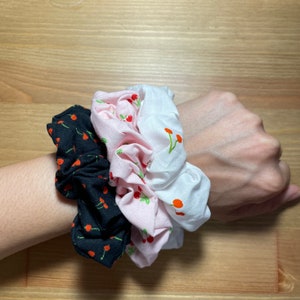 Könnte beinhalten: Drei Stoff-Haargummis mit Kirschendruck. Ein Haargummi ist schwarz, eines ist rosa und eines ist weiß.
