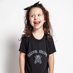 May include: A young girl wearing a black t-shirt with a white graphic of a fly and the text "SUPA DUPA" in a varsity style font. She is wearing a black headband and purple pants.