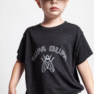 May include: A young boy wearing a black and grey t-shirt with a white fly graphic and the text "SUPA DUPA" in a black and white arch. He is also wearing a black and grey trucker hat with the Hurley logo.