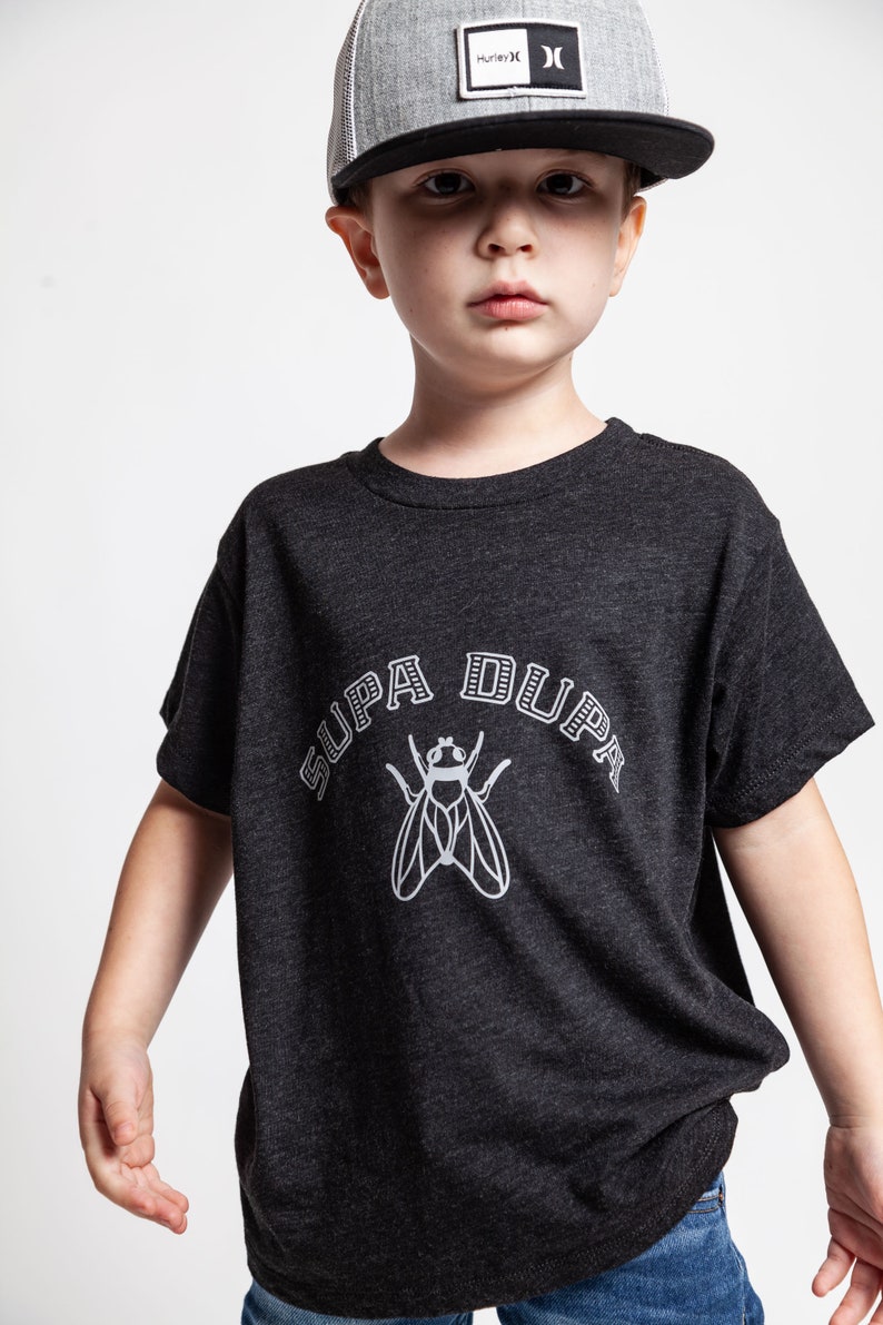 May include: A young boy wearing a black and grey t-shirt with a white fly graphic and the text "SUPA DUPA" in a black and white arch. He is also wearing a black and grey trucker hat with the Hurley logo.