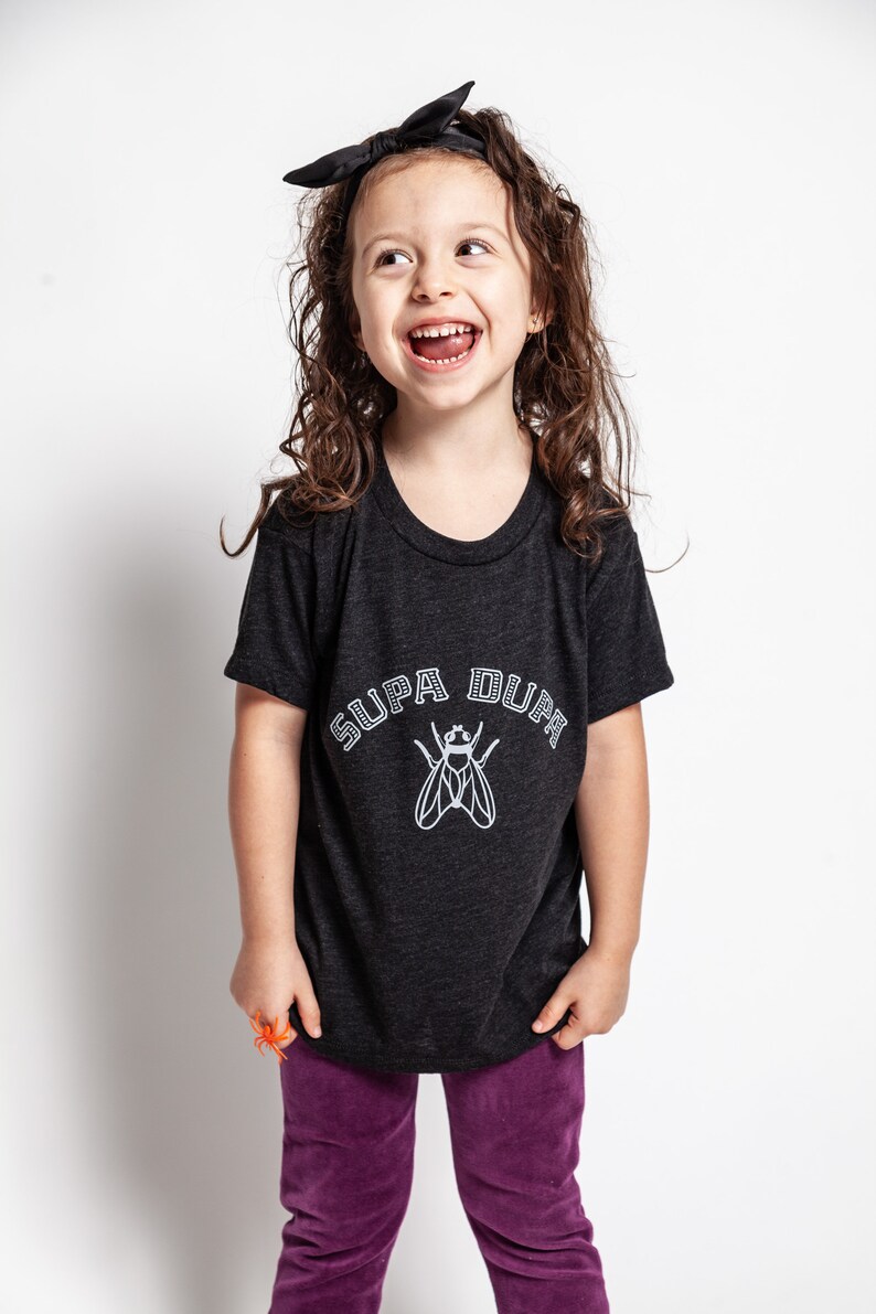 May include: A young girl wearing a black t-shirt with a white graphic of a fly and the text "SUPA DUPA" in a varsity style font. She is wearing a black headband and purple pants.