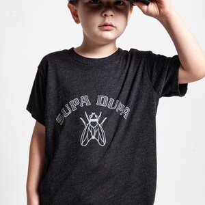 May include: A young person wearing a black t-shirt with a white graphic of a fly and the text "SUPA DUPA". The person is also wearing a gray and black Hurley brand baseball cap.