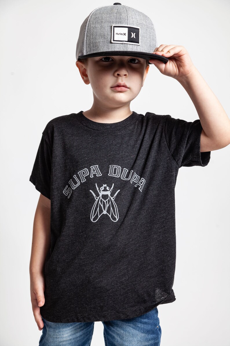 May include: A young person wearing a black t-shirt with a white graphic of a fly and the text "SUPA DUPA". The person is also wearing a gray and black Hurley brand baseball cap.