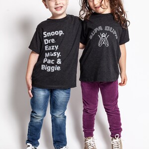 May include: Two children wearing black t-shirts with white text. The boy's shirt says "Snoop. Dre. Eazy. Missy. Pac & Biggie." The girl's shirt says "Supa Dupa" with a fly graphic. The boy is wearing blue jeans and black sneakers. The girl is wearing purple pants and leopard print sneakers.