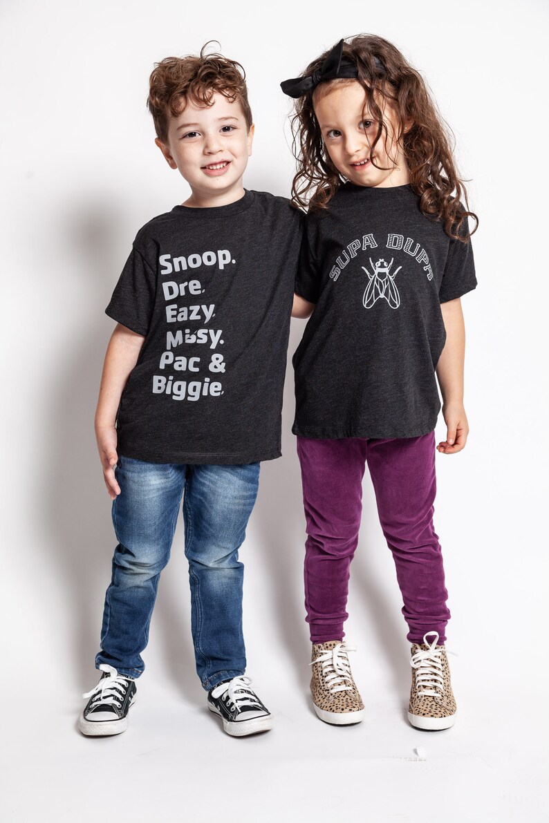 May include: Two children wearing black t-shirts with white text. The boy's shirt says "Snoop. Dre. Eazy. Missy. Pac & Biggie." The girl's shirt says "Supa Dupa" with a fly graphic. The boy is wearing blue jeans and black sneakers. The girl is wearing purple pants and leopard print sneakers.