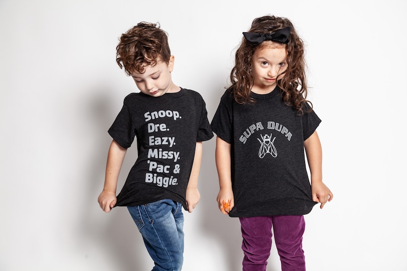 May include: Two children wearing black t-shirts with different designs. The boy's shirt has the names of several rappers printed on it, including Snoop, Dre, Eazy, Missy, 'Pac & Biggie. The girl's shirt has a fly with the words "SUPA DUPA" printed above it.