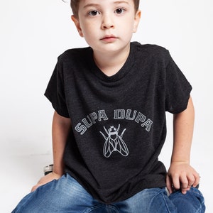 May include: A young boy wearing a dark grey t-shirt with a white graphic of a fly and the text "SUPA DUPA". He is wearing blue jeans and sitting on a white surface.