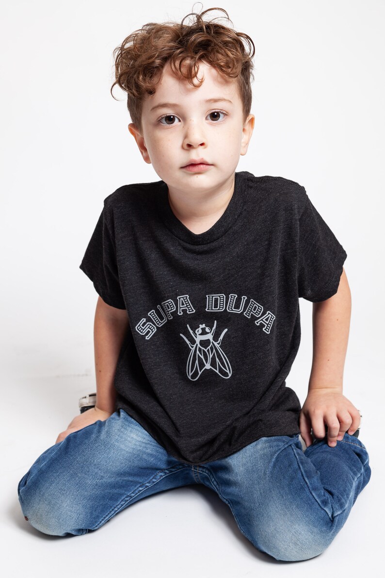 May include: A young boy wearing a dark grey t-shirt with a white graphic of a fly and the text "SUPA DUPA". He is wearing blue jeans and sitting on a white surface.