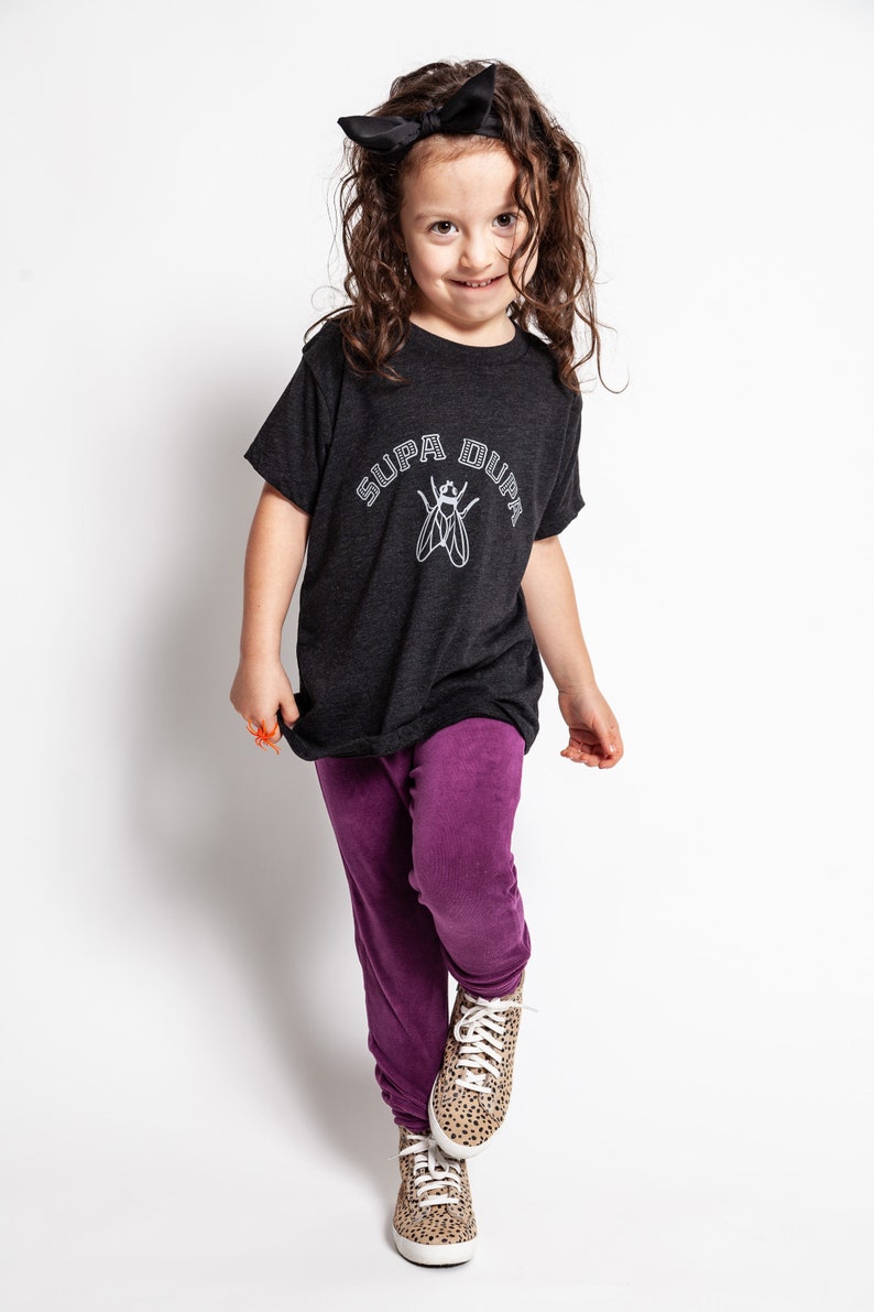 May include: A young person wearing a black t-shirt with the text "SUPA DUPA" and a fly graphic. They are also wearing purple pants and leopard print sneakers.