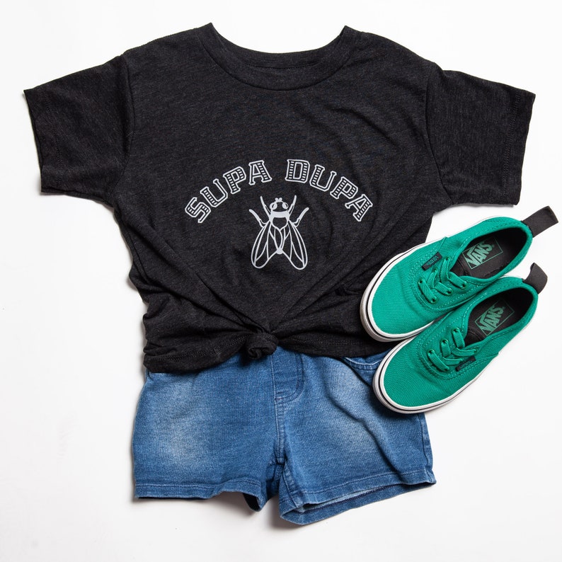 May include: A black t-shirt with the text "SUPA DUPA" in white lettering and a white fly graphic. The shirt is paired with blue denim shorts and green Vans sneakers.