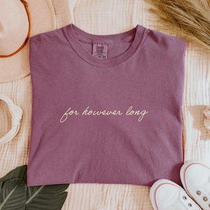May include: A mauve-colored t-shirt with the phrase "for however long" in cursive script. The shirt is a crew neck style and appears to be made of a soft material. The shirt is laying on a cream-colored surface, with white sneakers visible in the bottom right corner.