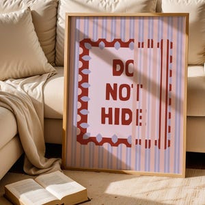Puede incluir: Una impresión artística enmarcada con las palabras "DO NOT HIDE" en letras rojas. La obra de arte presenta un fondo a rayas rosas y blancas con un borde rojo. El marco es de madera clara.